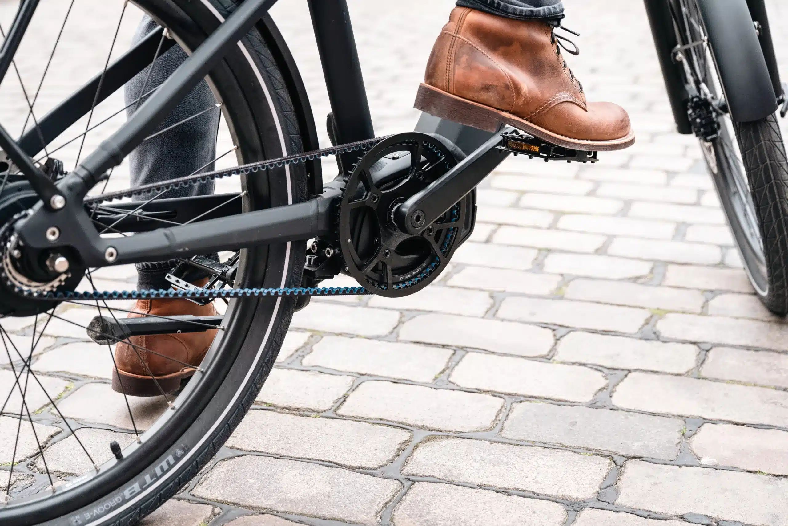 Detailaufnahme des Antriebs eines HNF XD4 E-Bikes mit schwarzem Gates Carbon Drive Riemen, Kettenblatt und Pedal in Lederstiefel. Das Fahrrad steht auf einem Kopfsteinpflaster.