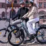 Zwei Personen fahren Kemoway City E-Bike K5 auf einem Platz in der Stadt, beide tragen Helme und lächeln