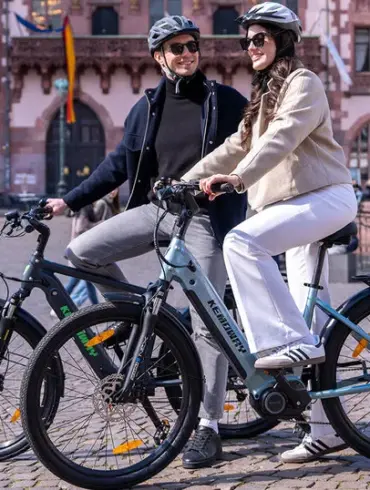 Zwei Personen fahren Kemoway City E-Bike K5 auf einem Platz in der Stadt, beide tragen Helme und lächeln