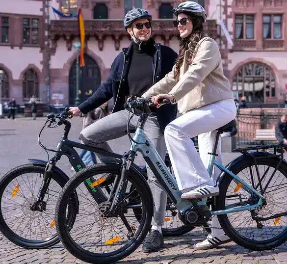 Zwei Personen fahren Kemoway City E-Bike K5 auf einem Platz in der Stadt, beide tragen Helme und lächeln