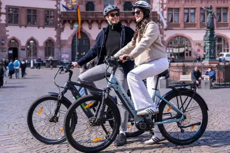 Zwei Personen fahren Kemoway City E-Bike K5 auf einem Platz in der Stadt, beide tragen Helme und lächeln