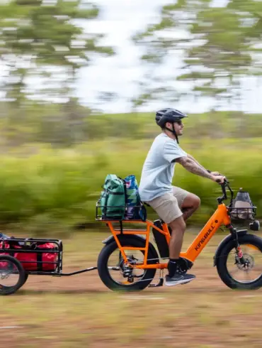 Ein Mann auf einem orangefarbenen Kingbull Dual Battery Voyager 2.0 E-Bike mit Helm und Sonnenbrille, das einen Anhänger mit Gepäck zieht, während er auf einem Feldweg fährt.