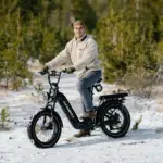 Ein Mann sitzt auf einem schwarzen Kingbull Jumper Go 20 Elektro-Geländebike mit dicken Reifen und vollgefedertem Rahmen. Er trägt eine beige Fleecejacke und eine Mütze und fährt auf verschneitem Untergrund, umgeben von Bäumen.