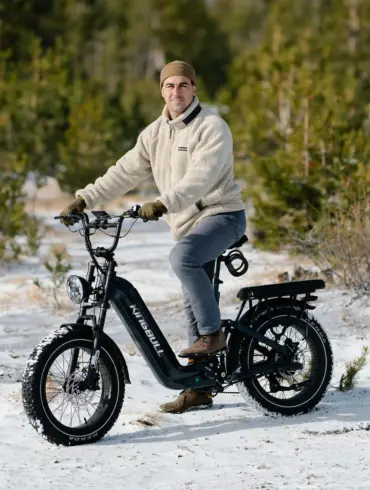 Ein Mann sitzt auf einem schwarzen Kingbull Jumper Go 20 Elektro-Geländebike mit dicken Reifen und vollgefedertem Rahmen. Er trägt eine beige Fleecejacke und eine Mütze und fährt auf verschneitem Untergrund, umgeben von Bäumen.