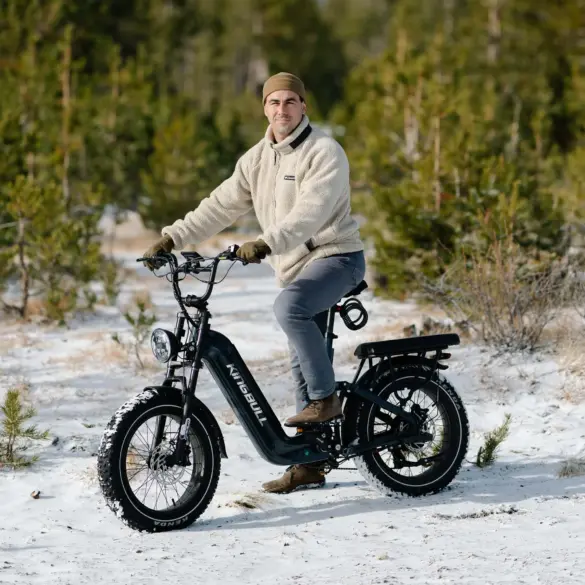 Ein Mann sitzt auf einem schwarzen Kingbull Jumper Go 20 Elektro-Geländebike mit dicken Reifen und vollgefedertem Rahmen. Er trägt eine beige Fleecejacke und eine Mütze und fährt auf verschneitem Untergrund, umgeben von Bäumen.