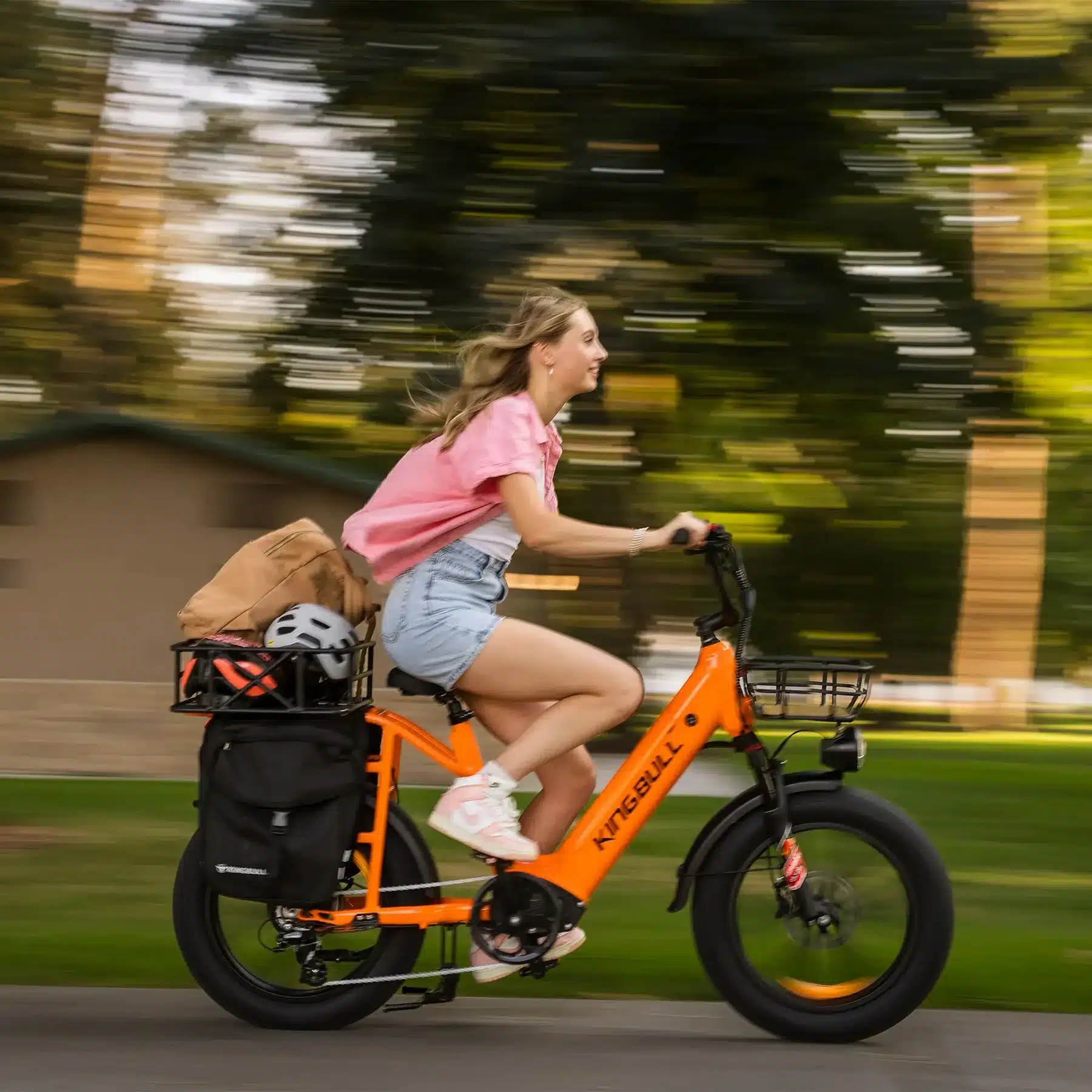 Orangefarbenes Kingbull Voyager 2.0 E-Bike mit breiten Reifen, Vorderrad- und Heckträger, das von einer Frau mit Helm und Einkaufstaschen gefahren wird, wobei die Umgebung durch Bewegungsunschärfe dargestellt wird.