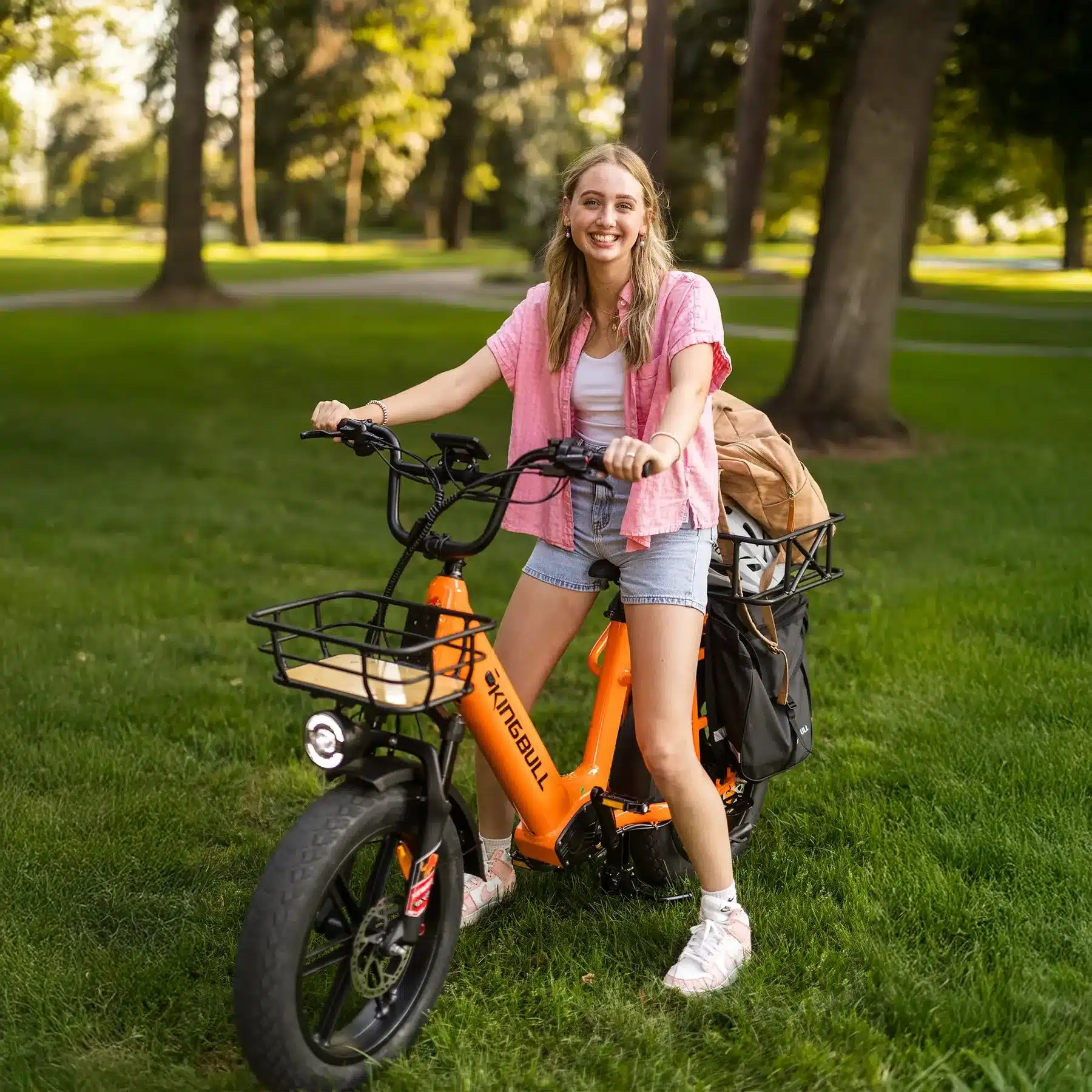 Junge Frau mit lächelndem Gesicht und einem orangefarbenen Kingbull Voyager 2.0 E-Bike mit Gepäck, das in einem Park auf einer grünen Wiese steht.