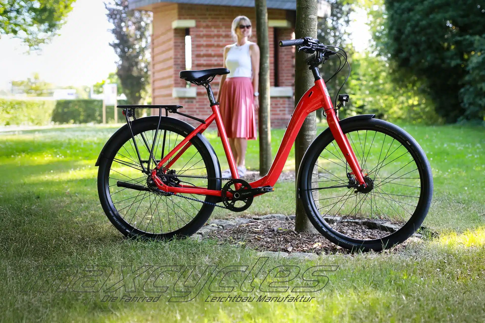 Rotes Maxcycles Lite Step 2 Fahrrad mit tiefem Einstieg, auf einer Rasenfläche stehend, mit einer Frau im Hintergrund.