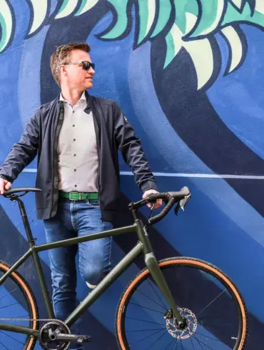 Ein Mann steht mit seinem dunkelgrünen Maxcycles Gravel Lite Fahrrad vor einer bunten Wand mit einem Wellenmuster.