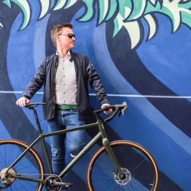 Ein Mann steht mit seinem dunkelgrünen Maxcycles Gravel Lite Fahrrad vor einer bunten Wand mit einem Wellenmuster.