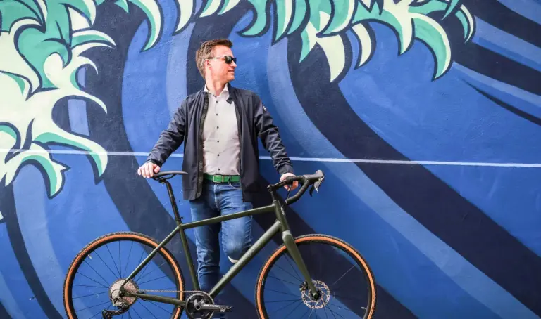 Ein Mann steht mit seinem dunkelgrünen Maxcycles Gravel Lite Fahrrad vor einer bunten Wand mit einem Wellenmuster.