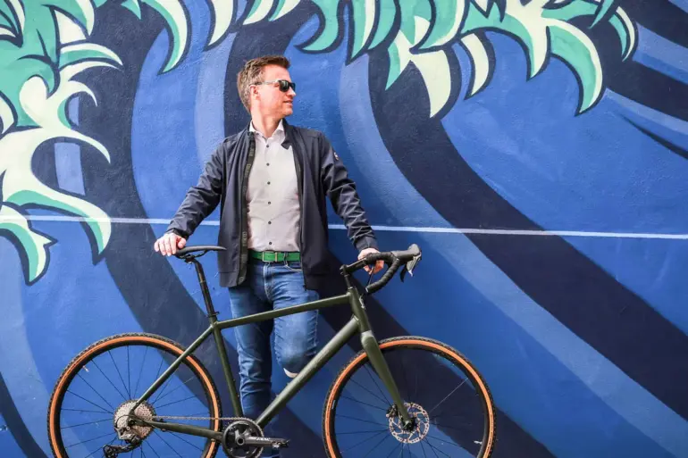Ein Mann steht mit seinem dunkelgrünen Maxcycles Gravel Lite Fahrrad vor einer bunten Wand mit einem Wellenmuster.