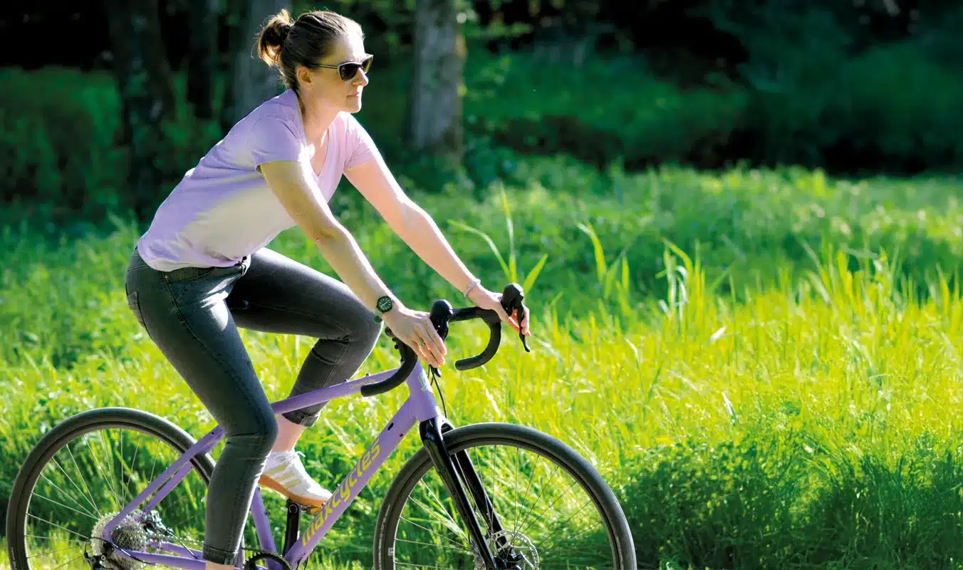 Frau mit Sonnenbrille fährt ein lila Maxcycles Gravel Lite Fahrrad auf einem Feldweg, umgeben von grüner Vegetation.