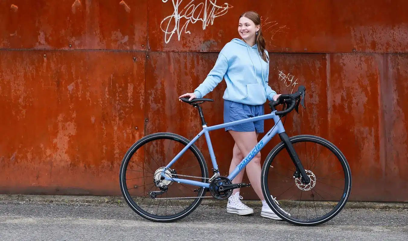 Frau mit einem blauen Maxcycles Gravel Lite Fahrrad vor einer rostigen Wand.