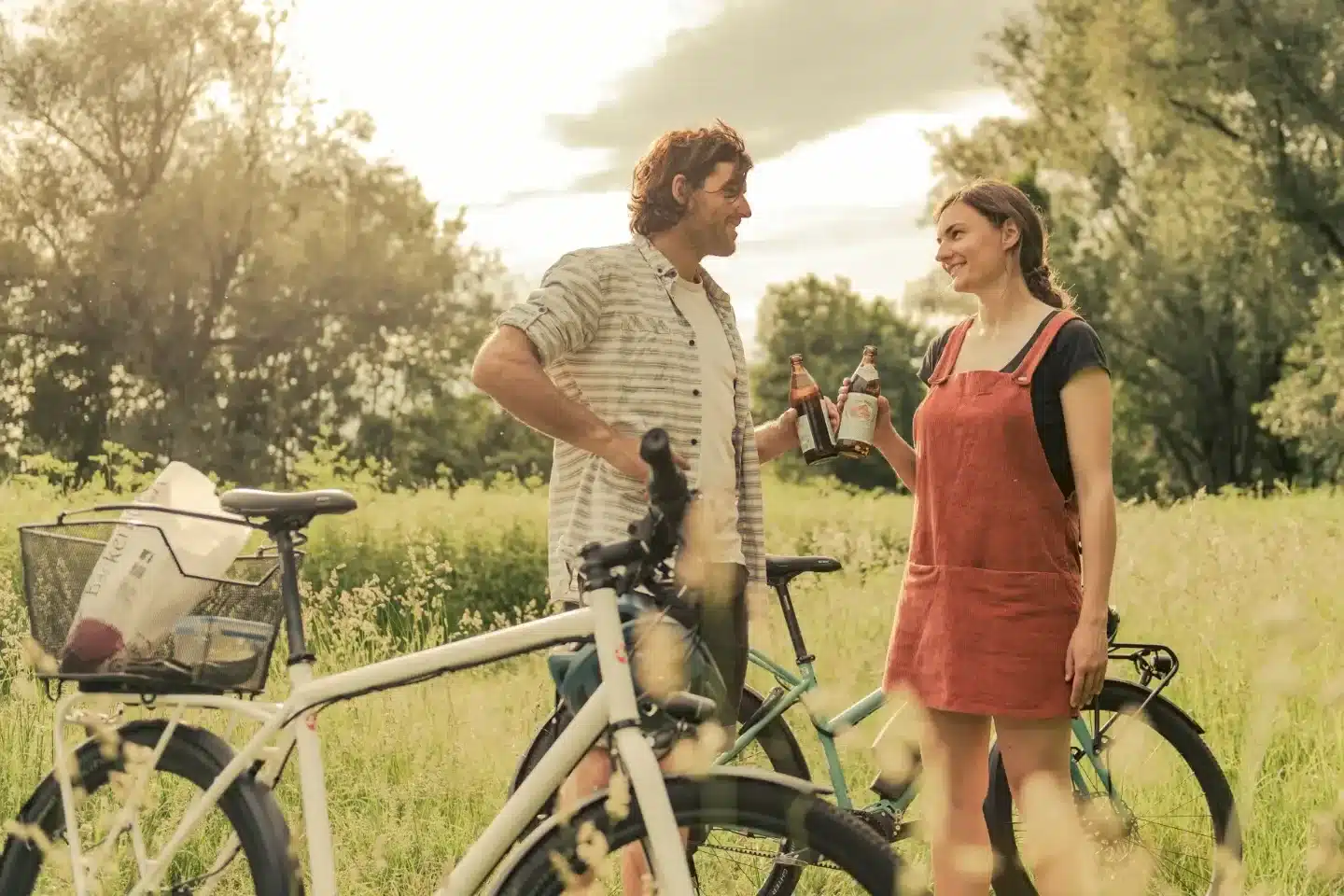 Mann und Frau mit Fahrrädern im Freien, sie halten Bierflaschen und unterhalten sich.