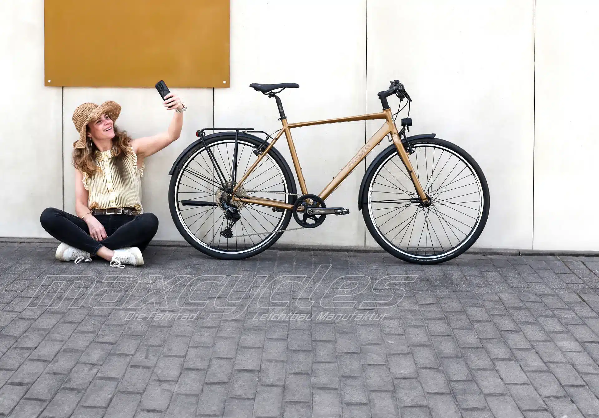 Moderne Frau mit Hut macht ein Selfie vor einem sportlichen Fahrrad neben einer weißen Wand.