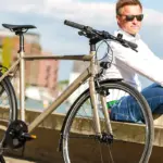 Nahaufnahme eines "Monza 2" Fahrrads in Beige, das neben einem Mann sitzt, der eine weiße Bluse und Jeans trägt, mit einer urbanen Kulisse im Hintergrund.