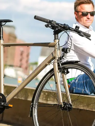 Nahaufnahme eines "Monza 2" Fahrrads in Beige, das neben einem Mann sitzt, der eine weiße Bluse und Jeans trägt, mit einer urbanen Kulisse im Hintergrund.