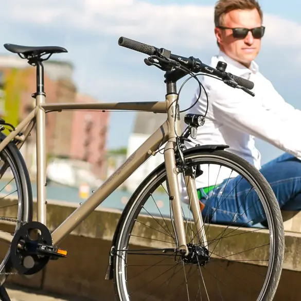 Nahaufnahme eines "Monza 2" Fahrrads in Beige, das neben einem Mann sitzt, der eine weiße Bluse und Jeans trägt, mit einer urbanen Kulisse im Hintergrund.