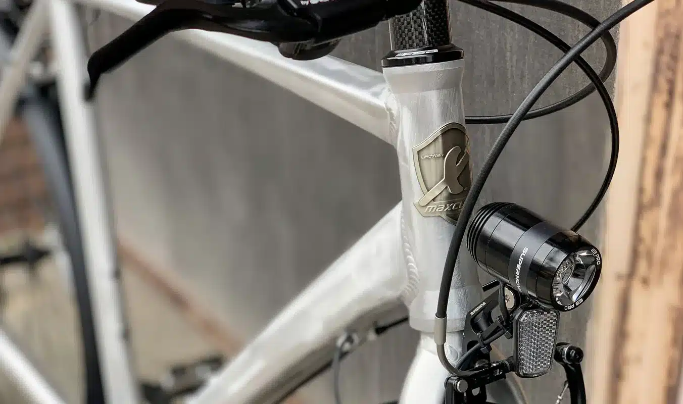 Nahaufnahme des hellsilbernen Aluminiumrahmens des Maxcycles Monza 2 Fahrrads. Das Bild zeigt Details wie das Markenlogo, den Gabelschaft und ein schwarzes Supernova E3 Pro Frontlicht mit Reflektor.