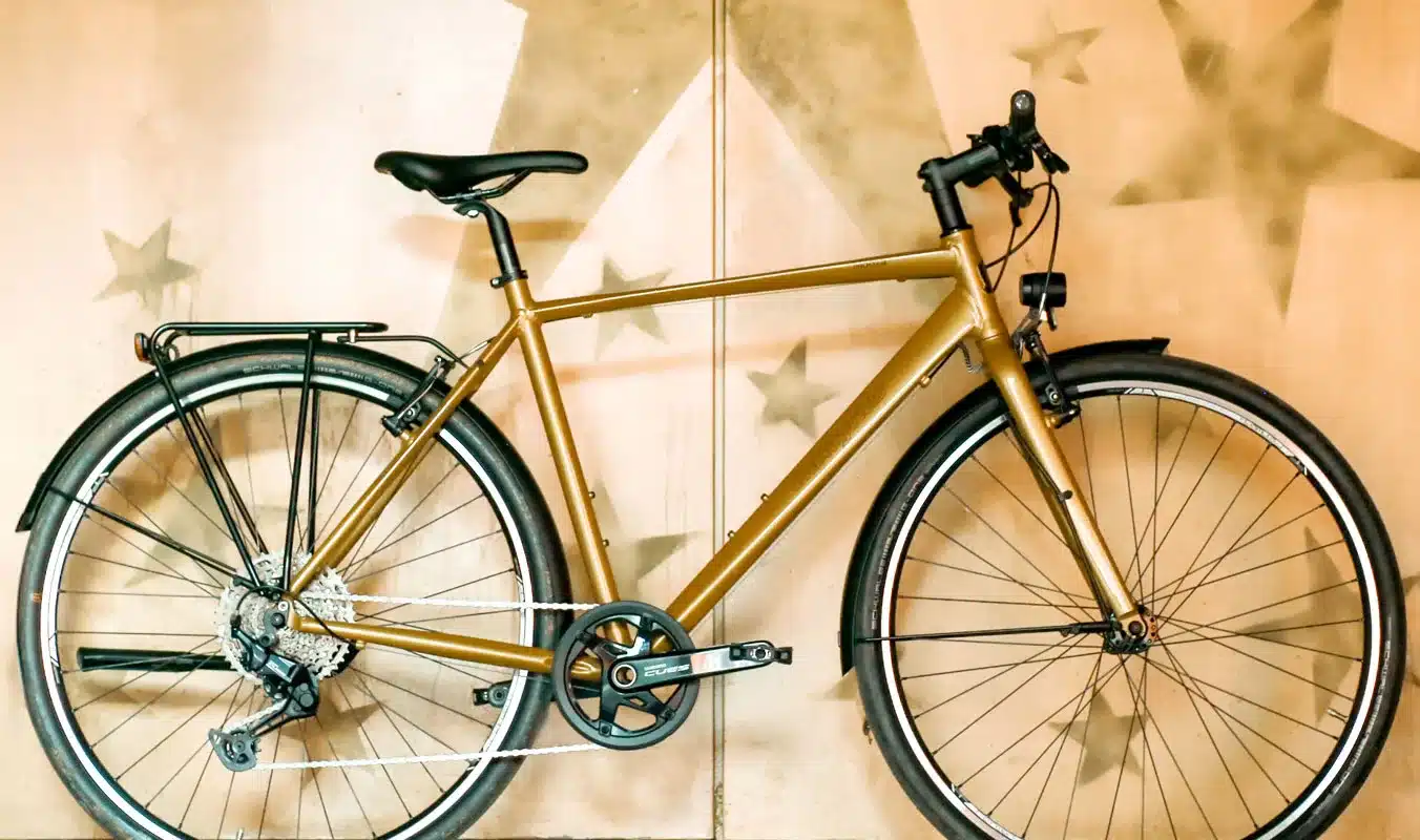 Seitliche Ansicht eines goldbraunen Monza 2 Fahrrads mit voll ausgestatteter Alltags- und Touring-Hardware wie Gepäckträger, Schutzblechen und integrierter Beleuchtung.