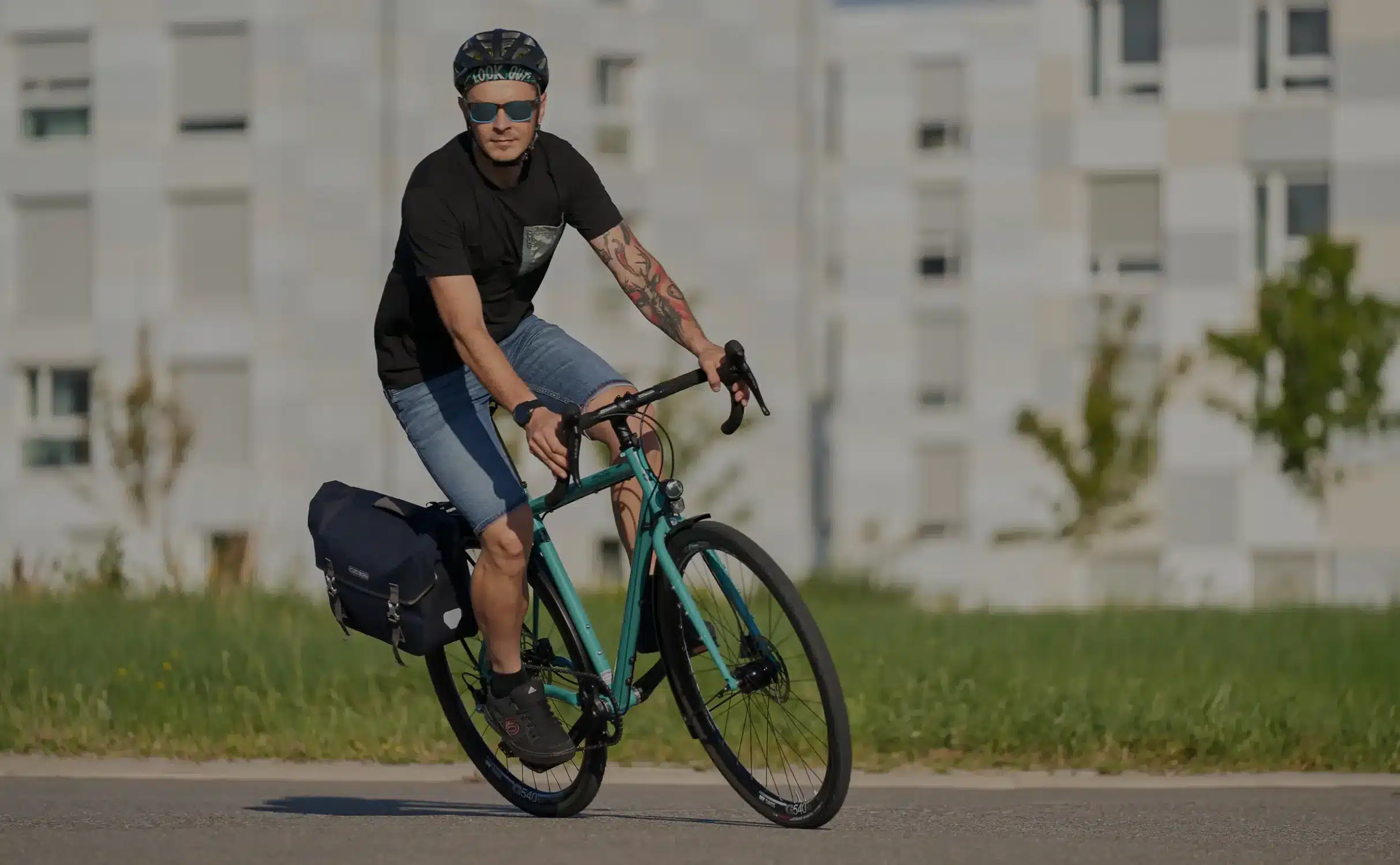 Ein Mann in Jeans und schwarzem T-Shirt fährt ein mintgrünes Tout Terrain Nolita Fahrrad mit Gepäcktasche am Hinterrad auf einer asphaltierten Straße.
