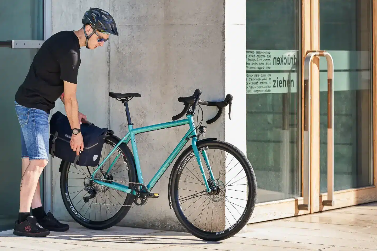 Mann mit Helm und Jeansshorts befestigt eine schwarze Ortlieb-Gepäcktasche an der Hinterradaufnahme eines türkisfarbenen Tout Terrain Nolita Allroad-Fahrrads, das neben einer Betonwand und einer Glastür steht.
