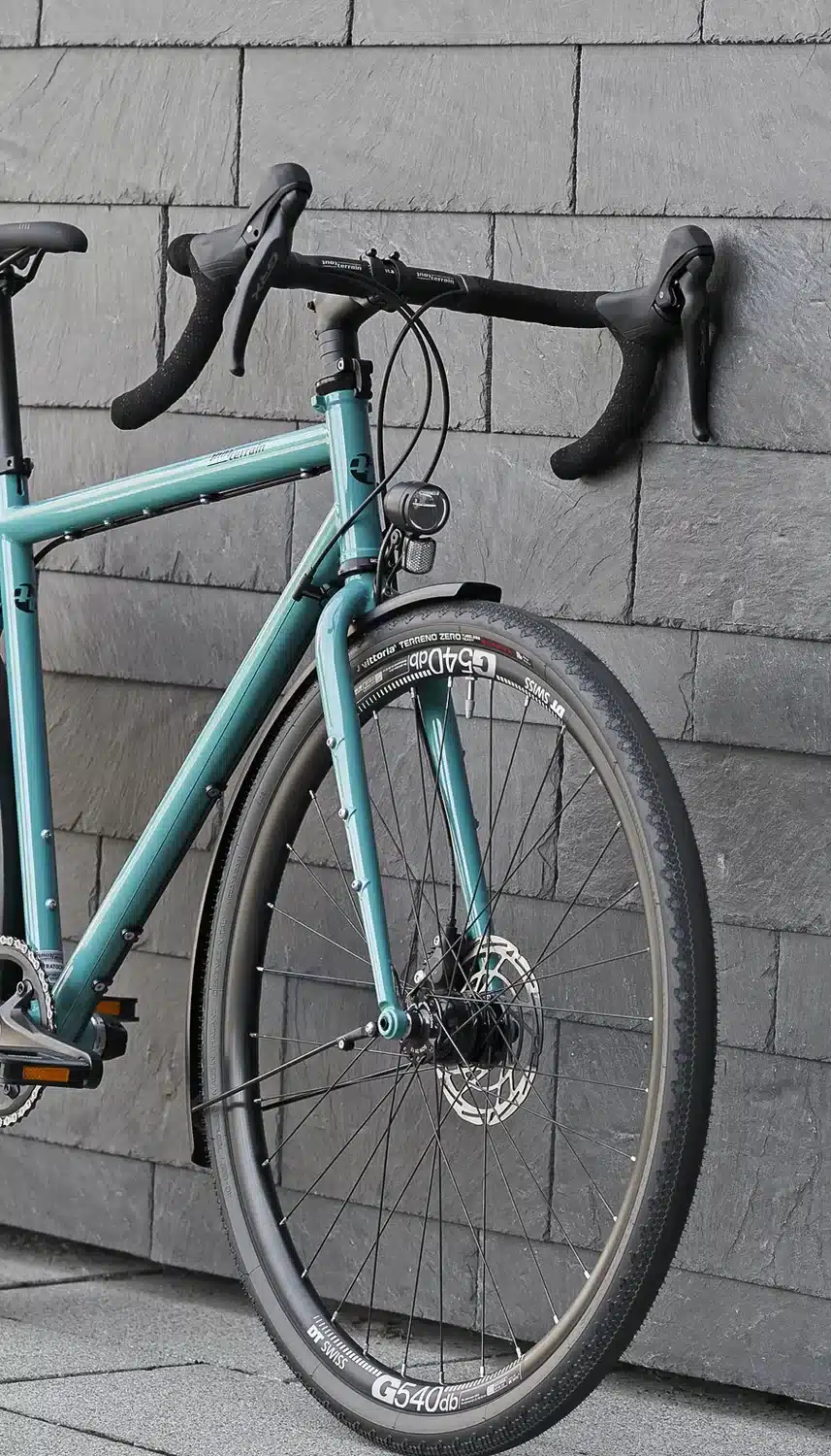 Teilansicht eines tealfarbenen Tout Terrain Nolita Fahrrads, das vor einer grauen Schieferwand steht, Fokus auf Vordergabel, Rad und Lenker.