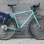 Profilansicht eines helltürkisen Tout Terrain Nolita Fahrrads mit einer dunkelblauen Ortlieb Gepäcktasche, das an einer grauen Steinwand steht.