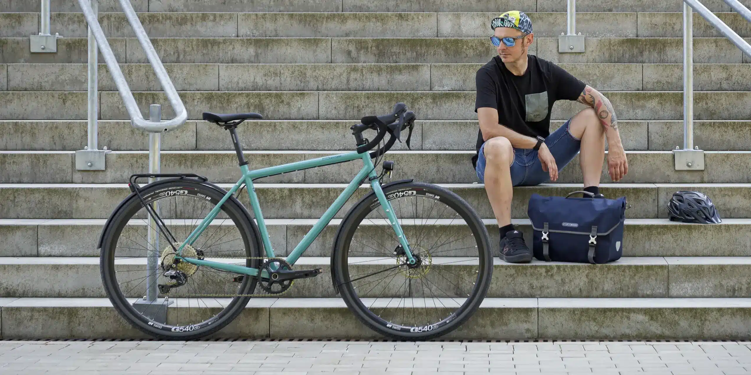 Ein Mann sitzt entspannt auf einer Steintreppe neben einem mintgrünen Tout Terrain Nolita Fahrrad. Daneben liegen eine Tasche und ein Helm.