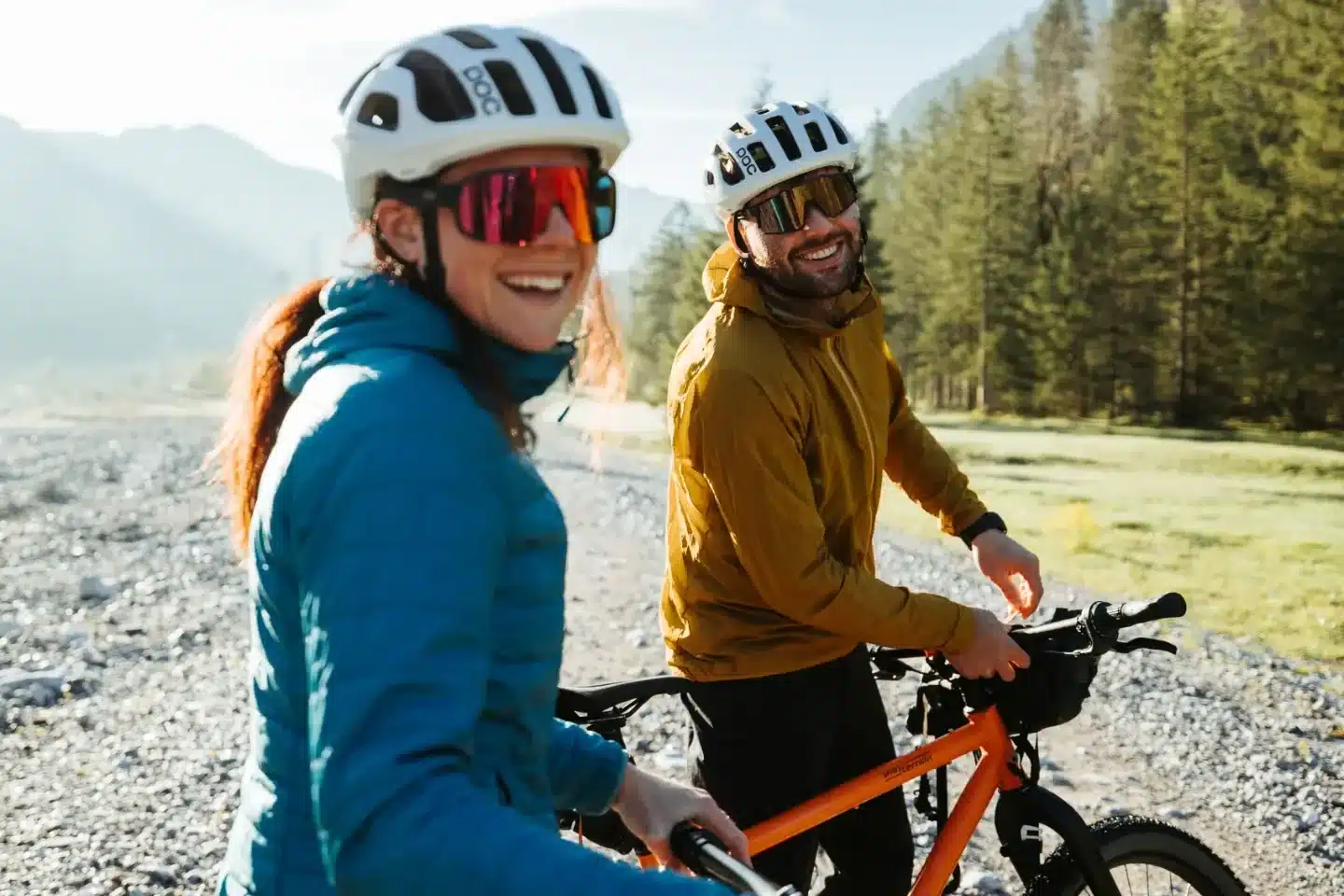 Ein Mann und eine Frau mit Fahrradhelmen und Sonnenbrillen stehen lächelnd auf einem Schotterweg im Gebirge, bereit für eine Radtour.