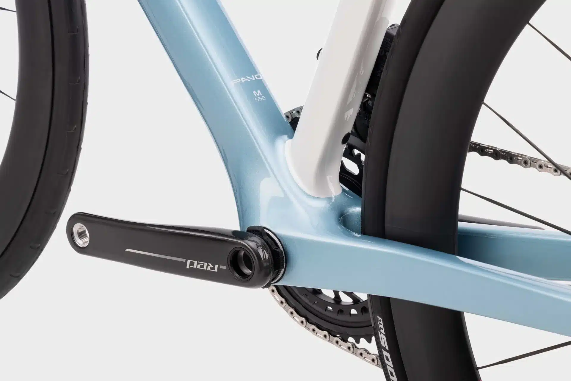 Detailaufnahme eines hellblauen Rennradrahmens mit der Aufschrift 'PAVO M 550'. Zu sehen ist das Tretlager, eine schwarze Kurbelgarnitur mit der Bezeichnung 'RED' und ein Teil des Kettenblatts sowie die Fahrradkette. Ein Reifen ist im Hintergrund sichtbar.