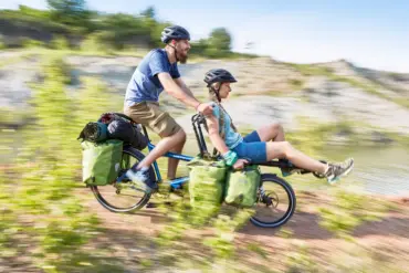 Zwei Personen, Mann und Frau, fahren auf einem blauen Hase Pino Tandem durch eine ländliche, hügelige Landschaft, beladen mit Gepäcktaschen und Schlafsäcken.
