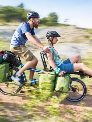 Zwei Personen, Mann und Frau, fahren auf einem blauen Hase Pino Tandem durch eine ländliche, hügelige Landschaft, beladen mit Gepäcktaschen und Schlafsäcken.