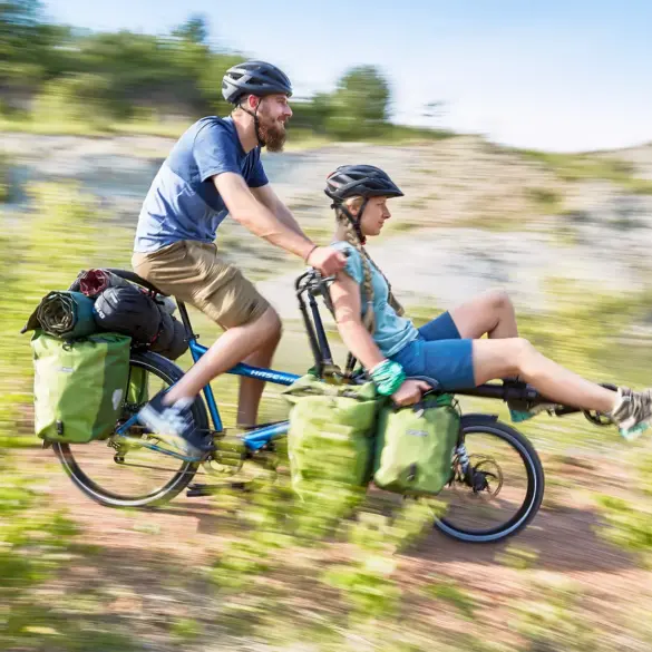 Zwei Personen, Mann und Frau, fahren auf einem blauen Hase Pino Tandem durch eine ländliche, hügelige Landschaft, beladen mit Gepäcktaschen und Schlafsäcken.