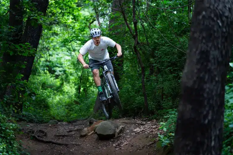 Ein Mountainbiker mit Helm und weißem T-Shirt fährt auf einem Hardtail-Bike einen felsigen und wurzeligen Waldweg entlang. Die Reifen sind breit und das Fahrrad scheint gut für anspruchsvolles Gelände geeignet zu sein.