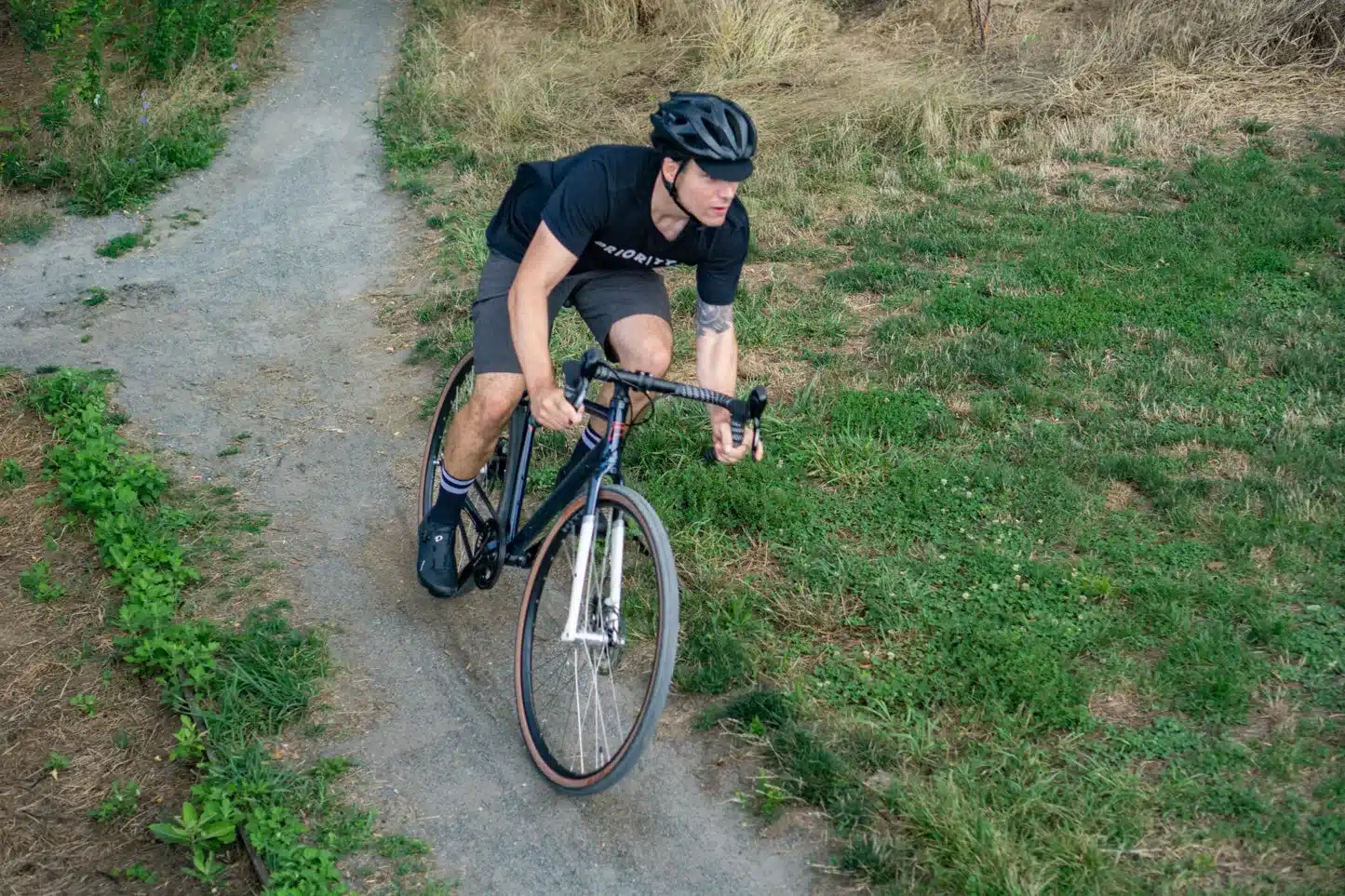 Mann mit Helm fährt Gravelbike auf einem Schotterweg, umgeben von Grünflächen und trockenem Gras.