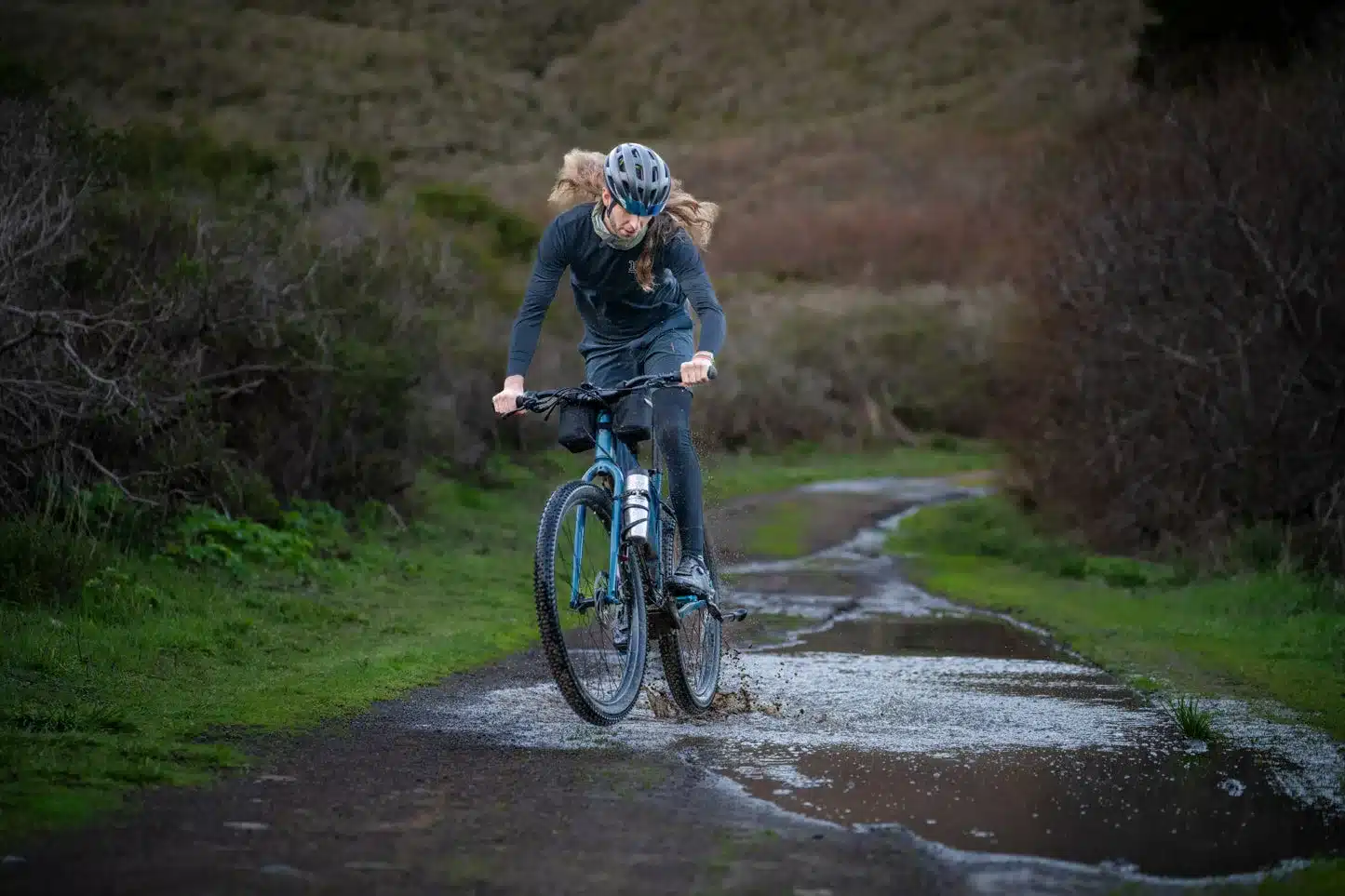 Ein Mann mit Helm und Brille fährt auf einem blauen Mountainbike durch eine tiefe Pfütze auf einem Feldweg im Freien.