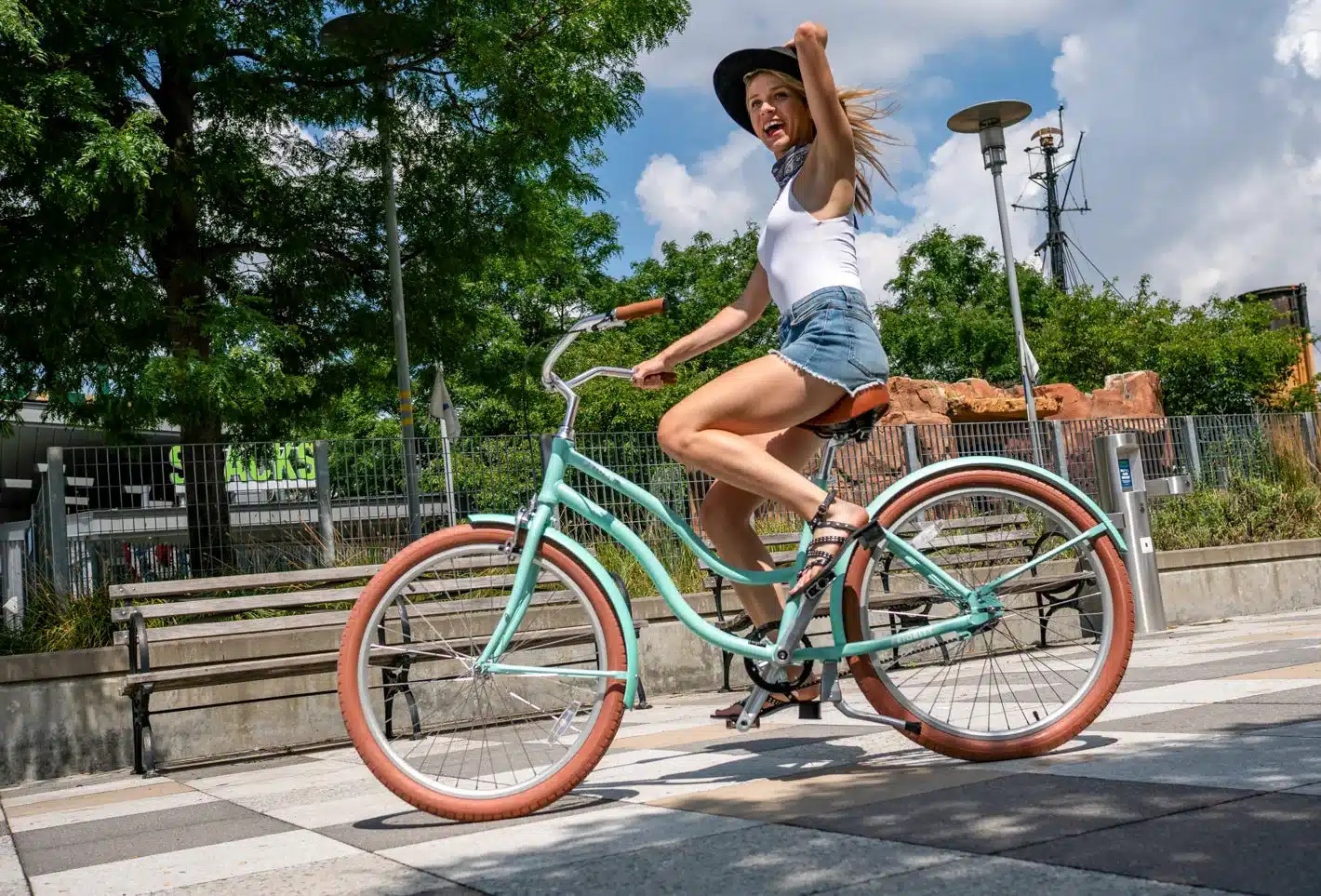 Eine Frau mit einem schwarzen Hut und einer weißen Weste fährt ein mintgrünes Fahrrad mit braunem Sattel und braunen Griffen auf einem Gehweg mit kariertem Muster.