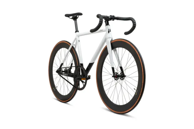 Studioaufnahme eines weißen Priority Joker Bahnrads mit schwarzen Lenkerbandern, Sattel und einer schwarzen Lackierung am Unterrohr und an der Gabelspitze. Das Rad hat tiefe schwarze Carbonfelgen mit braunen Reifenflanken und einen Gates CDX Carbon Drive Riemenantrieb.