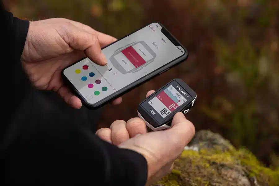 Person hält den SIGMA ROX 11.1 EVO GPS-Trainingcomputer und ein Smartphone mit geöffneter SIGMA RIDE App in der Hand