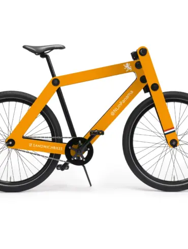 Ein oranges Sandwichbike WF1-S mit Holzrahmen und schwarzen Rädern steht auf weißem Hintergrund. Das Fahrrad hat eine auffällige orangefarbene Rahmenkonstruktion aus Holz und ist mit schwarzen Reifen und Felgen ausgestattet. Auf dem Oberrohr ist ein stilisiertes weißes Logo und der Schriftzug "@NLinPanama" angebracht, während am Rahmenende das "Sandwichbikes"-Logo zu sehen ist. Kleine farbige Streifen in Rot, Weiß und Blau zieren die Gabel.