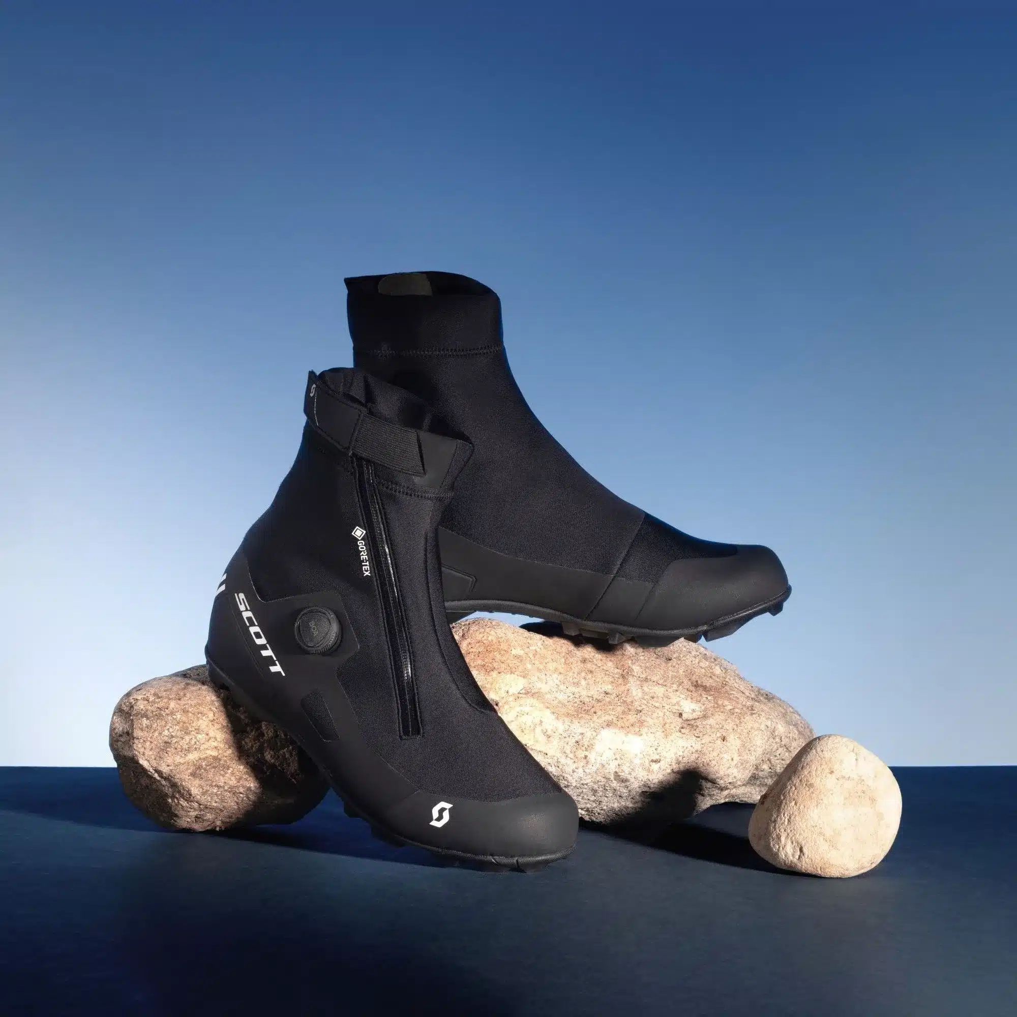 SCOTT MTB Heater Schuhe in Schwarz mit hohem Schaft und Reißverschluss, auf Steinen vor blauem Hintergrund platziert