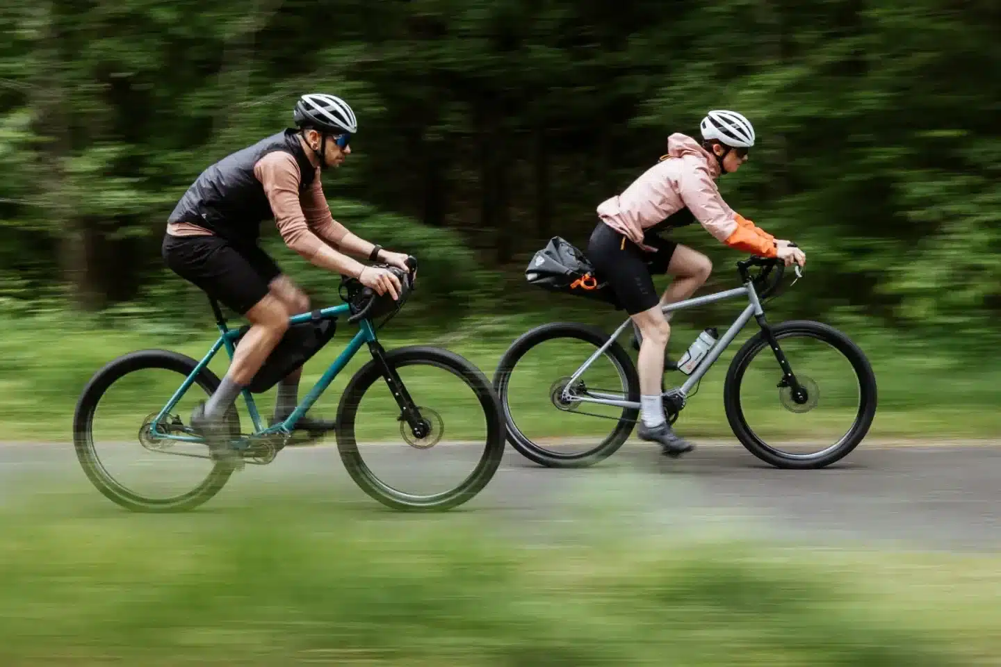 Zwei Radfahrer auf Scrambler Xplore GT II 28 Gravelbikes, mit Gepäcktaschen, fahren nebeneinander auf einer Straße durch einen grünen Wald.
