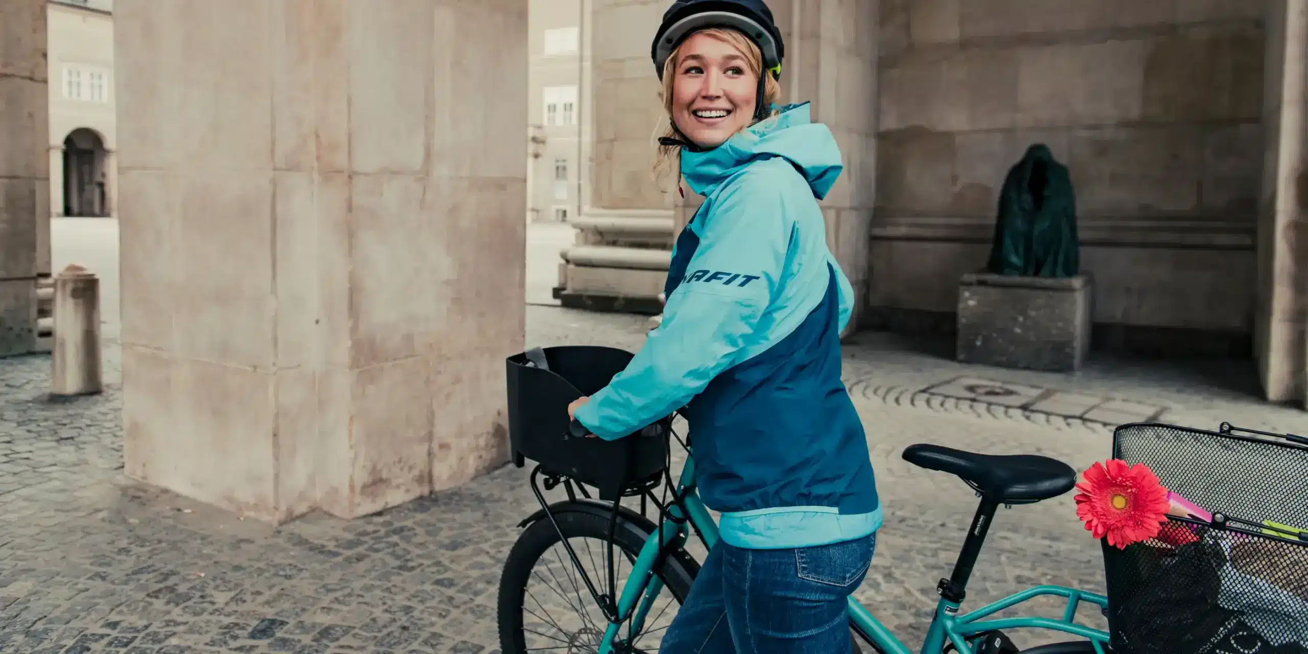 Lächelnde Frau mit blau-türkiser Jacke und Helm fährt ein türkisfarbenes Fahrrad mit zwei Körben durch einen steinernen Durchgang.