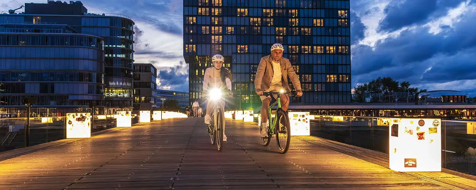 Zwei Radfahrer fahren bei Dämmerung mit eingeschalteter Sigma AURA 50 Frontlampe über eine moderne, beleuchtete Brücke vor urbaner Kulisse.