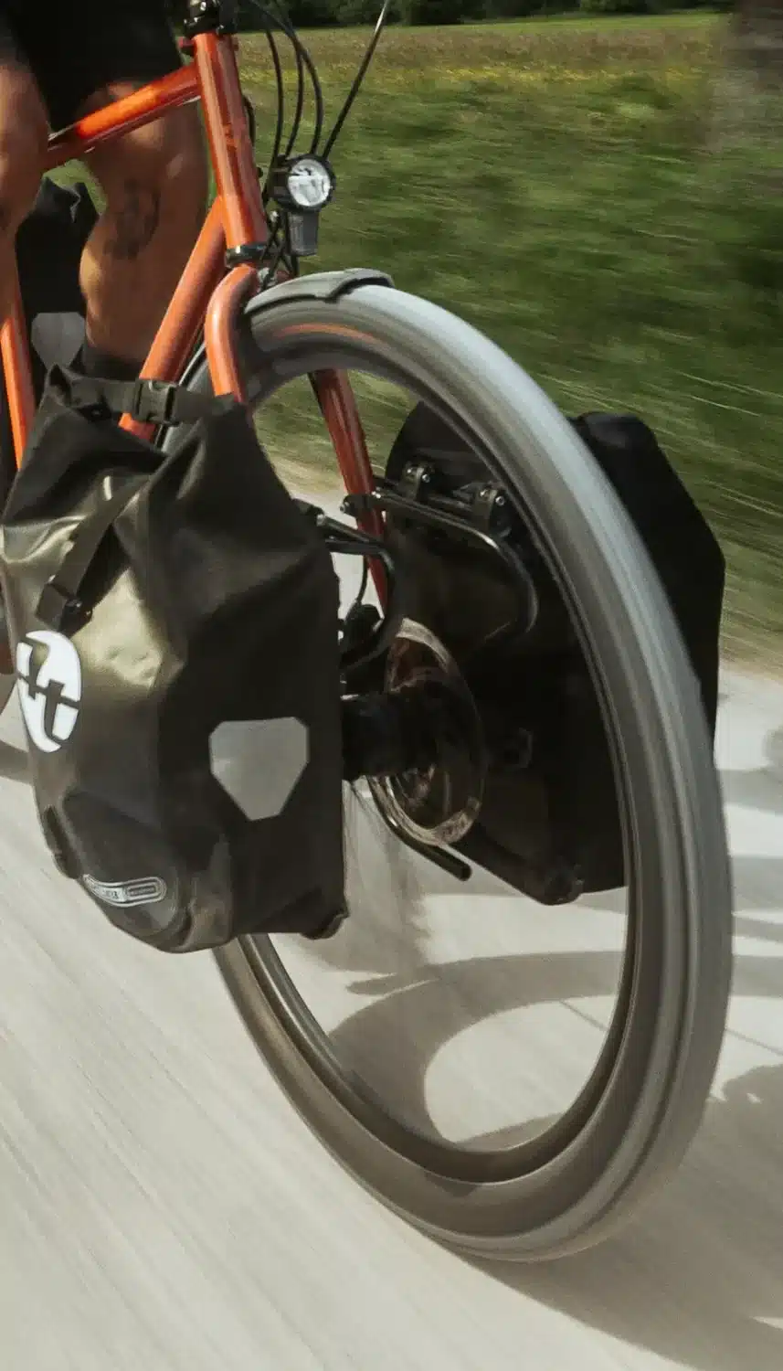 Nahaufnahme eines orangefarbenen Tout Terrain Silkroad Reiserads, das mit einer schwarzen Ortlieb Gepäcktasche am Vorderrad und einem Schutzblech ausgestattet ist, während es auf einem Feldweg fährt.