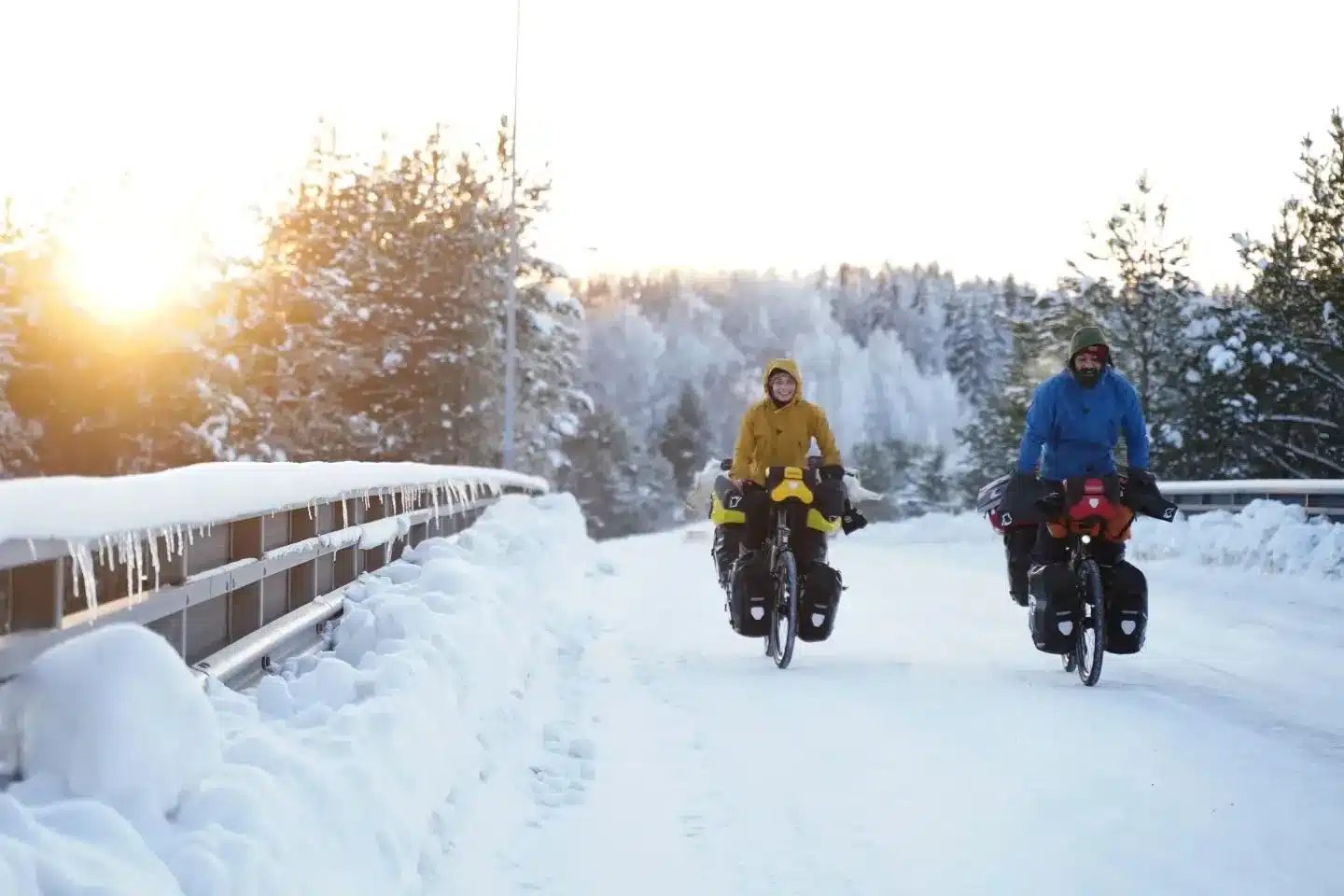 Zwei Radreisende fahren auf einer verschneiten Straße mit beladenen Fahrrädern und Winterkleidung.