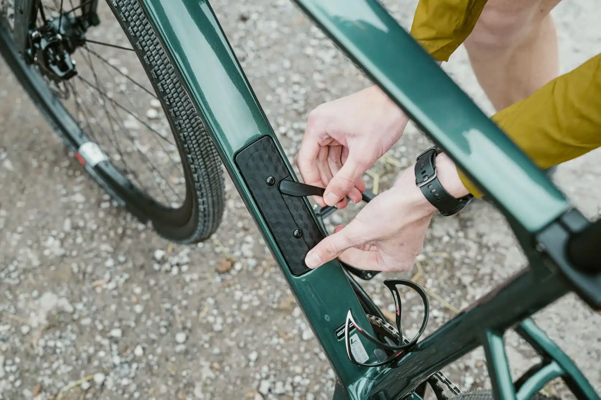 Detailaufnahme eines dunkelgrünen SIMPLON GRID :eLight E-Gravelbikes, bei der eine Person mit der Hand eine Klappe am Unterrohr öffnet, vermutlich um Zugang zur Batterie zu erhalten. Das Vorderrad des Fahrrads und ein Teil des Rahmens sind im Bild sichtbar.