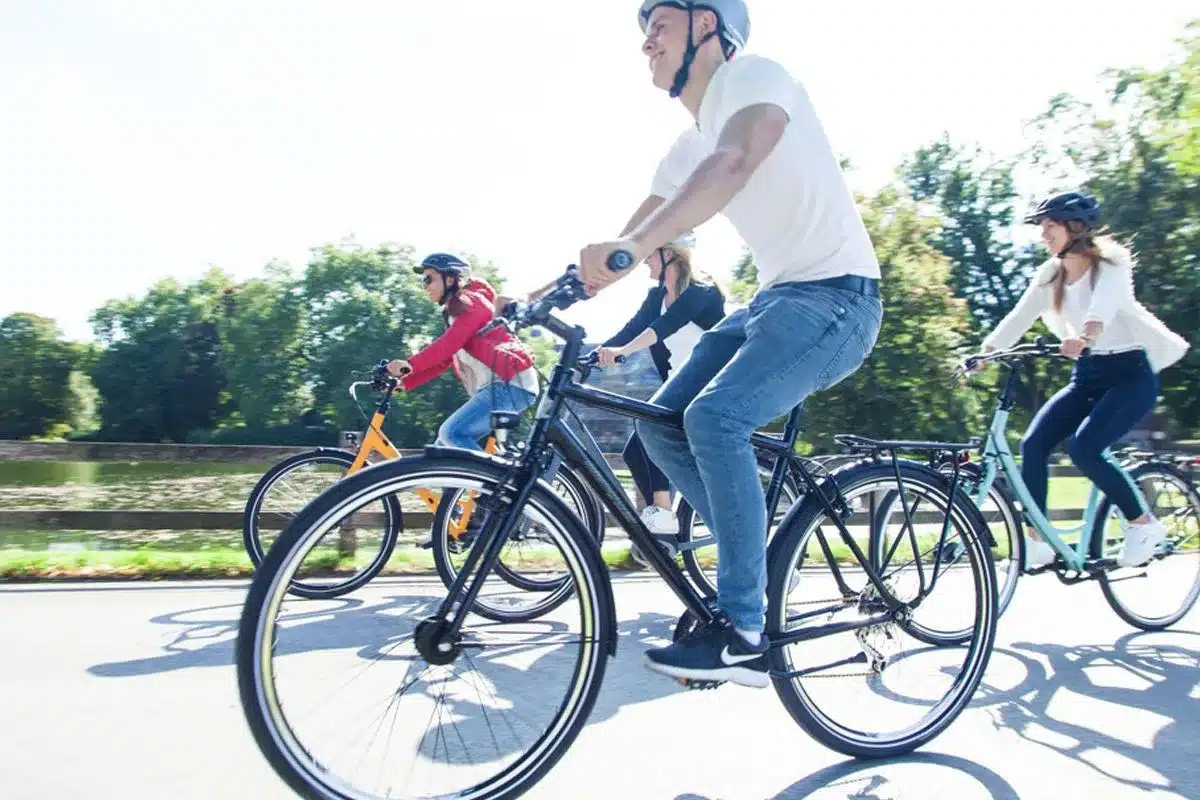 Vier Personen fahren Fahrrad, einige mit Helmen, auf einer asphaltierten Straße, mit Bäumen im Hintergrund